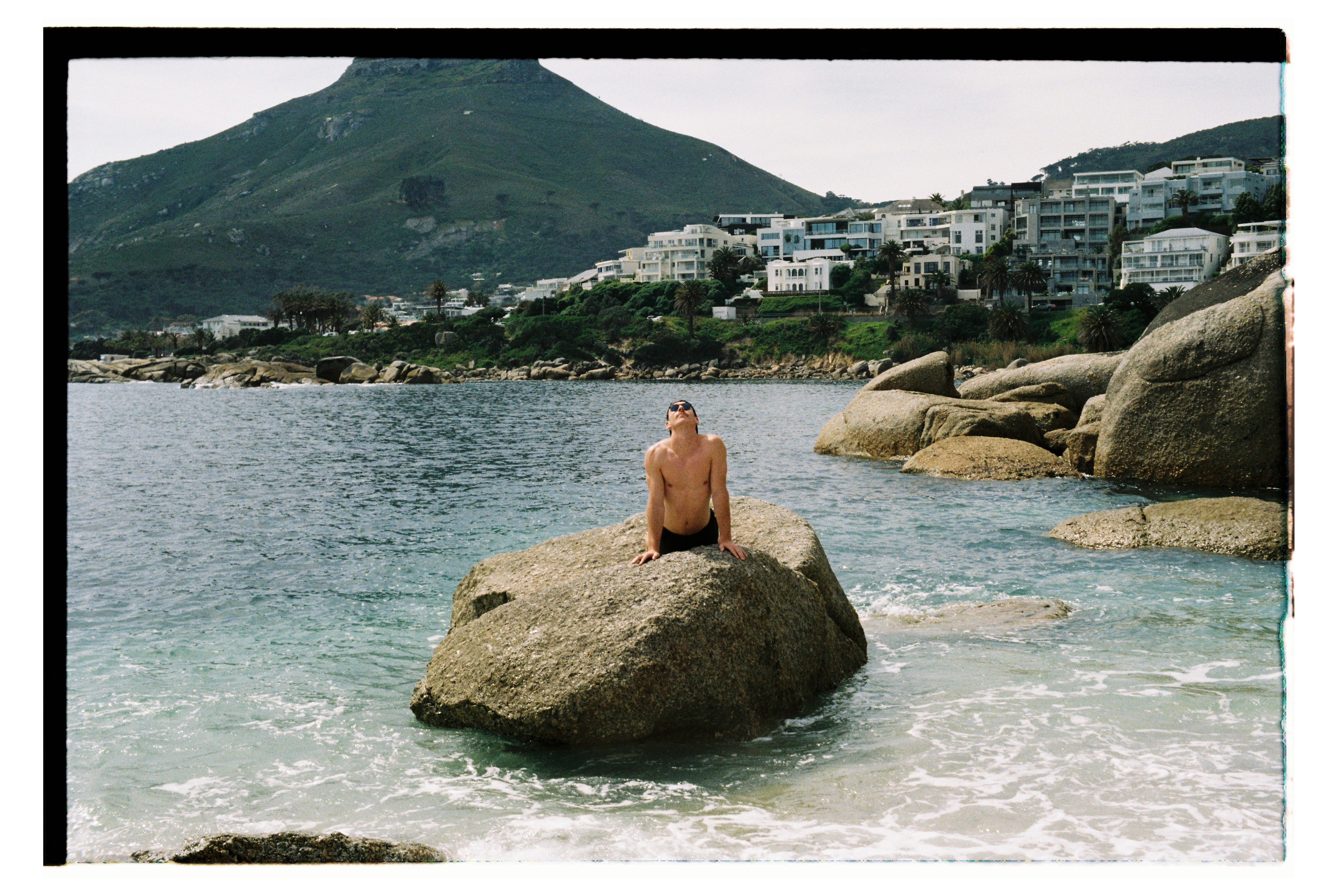 Man poses like a mermaid on a rock by the shore.